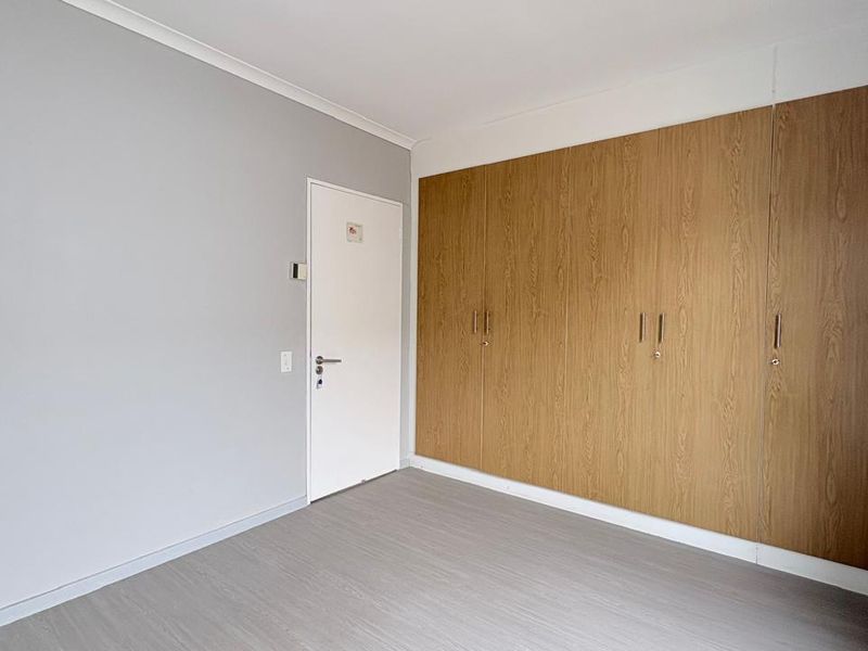 A clean, empty bedroom featuring light grey laminate flooring, neutral walls, and extensive built-in wooden cupboards providing ample storage space.
