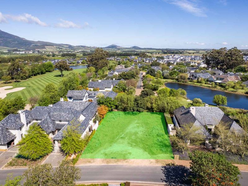 An aerial view of a vacant residential plot situated in a scenic estate, featuring surrounding greenery, water features, and established neighboring homes.