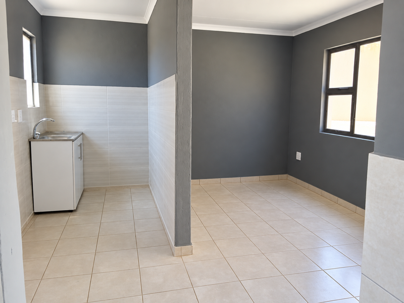An open-plan living and kitchen area featuring neutral tiled flooring, modern grey walls, and a compact kitchenette with a stainless steel sink.