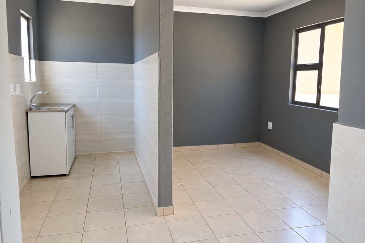 An open-plan living and kitchen area featuring neutral tiled flooring, modern grey walls, and a compact kitchenette with a stainless steel sink.