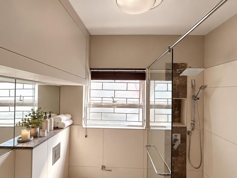Modern bathroom featuring a glass-enclosed shower with rain and handheld shower heads, built-in cabinetry, and neutral wall tiling.