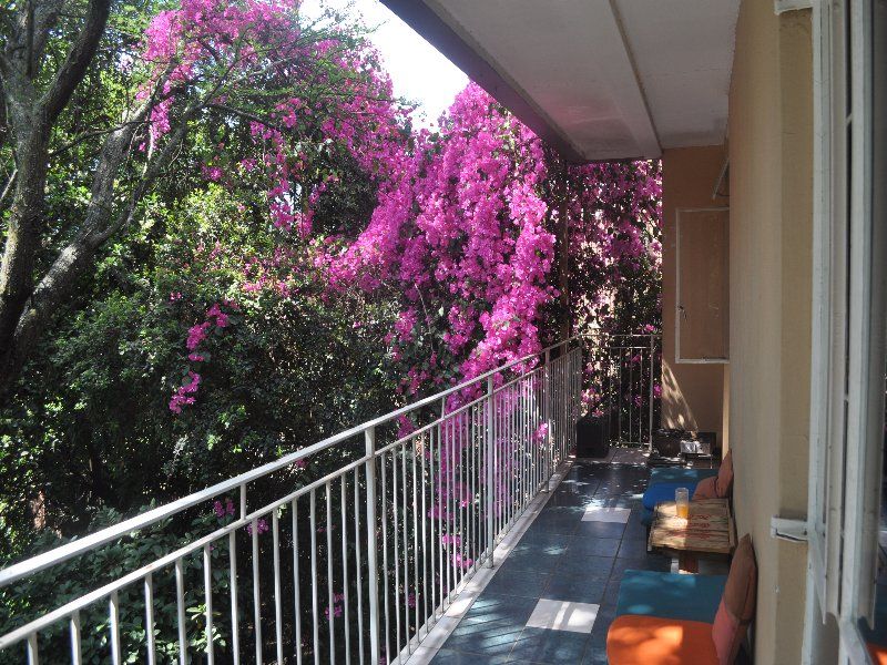 A covered balcony featuring tiled flooring and a metal balustrade, overlooking lush greenery and vibrant bougainvillea.
