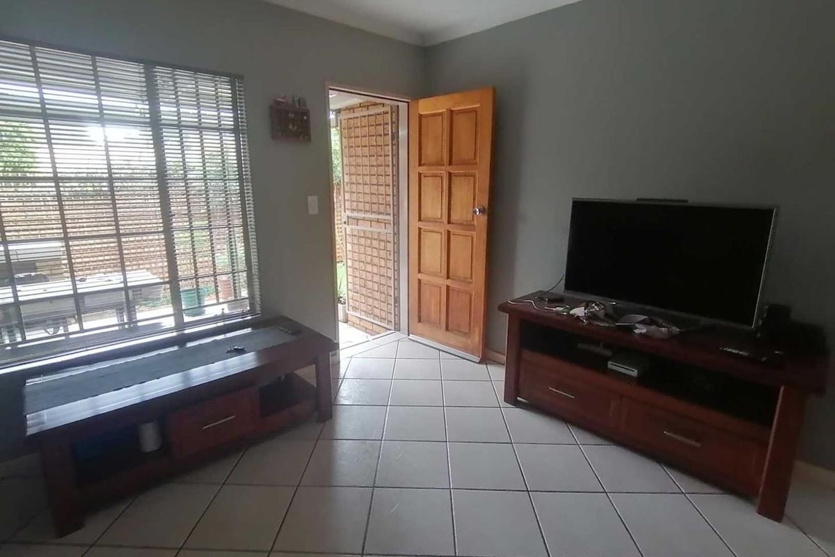 A living room featuring tiled flooring, a large window with blinds, and a wooden door leading to the exterior.