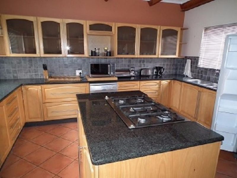 A functional kitchen featuring wooden cabinetry, granite countertops, a central island with a gas hob, and tiled flooring with exposed timber ceiling beams.