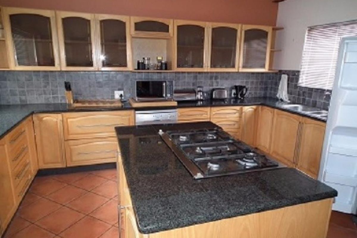 A functional kitchen featuring wooden cabinetry, granite countertops, a central island with a gas hob, and tiled flooring with exposed timber ceiling beams.
