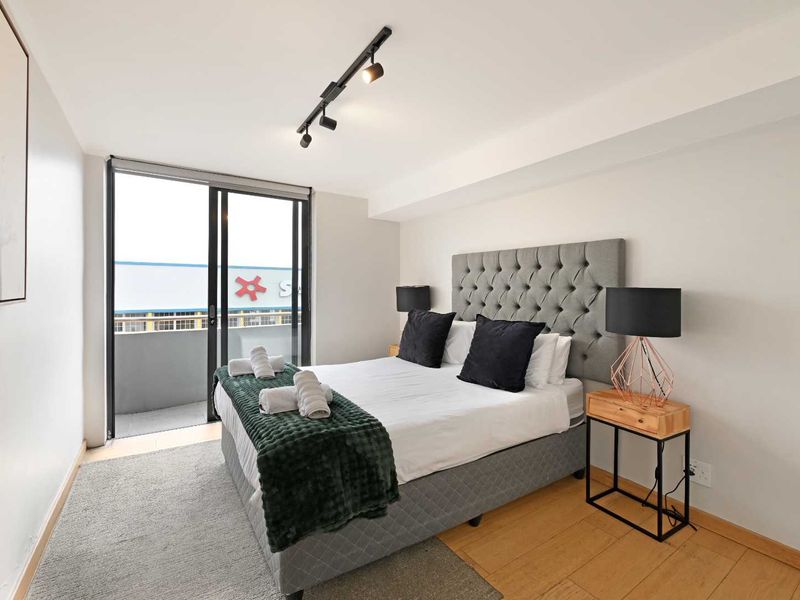 A well-lit bedroom featuring timber flooring, modern track lighting, and a sliding glass door providing access to a private balcony.