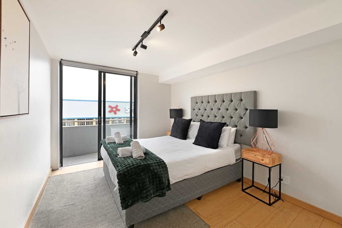 A well-lit bedroom featuring timber flooring, modern track lighting, and a sliding glass door providing access to a private balcony.