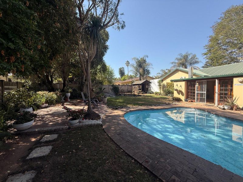 A spacious backyard featuring a swimming pool with brick coping, a paved patio area, and a well-established garden with mature trees.