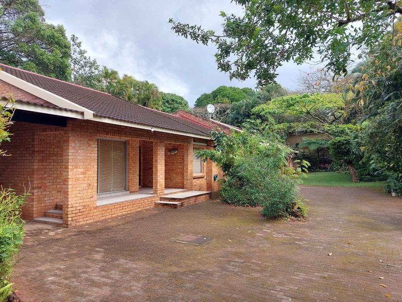 Rear view of a brick townhouse featuring a covered patio, paved driveway, and established garden surroundings.