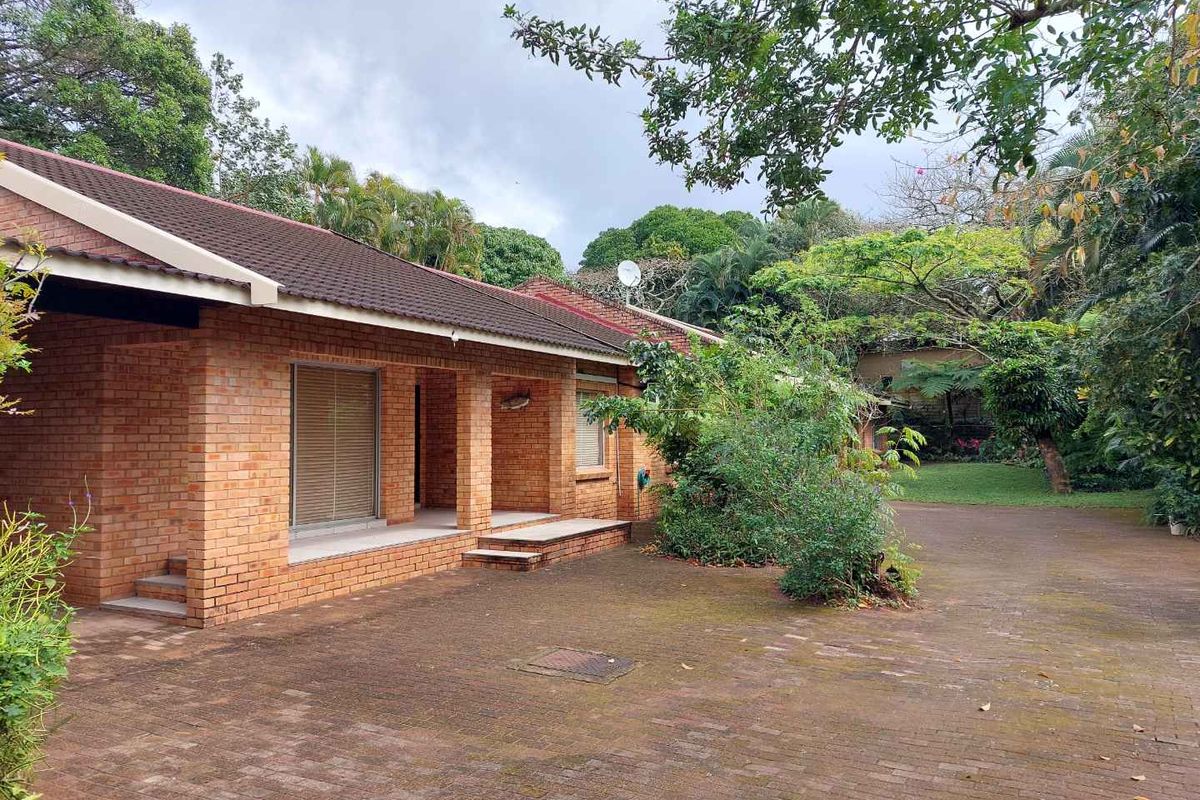 Rear view of a brick townhouse featuring a covered patio, paved driveway, and established garden surroundings.