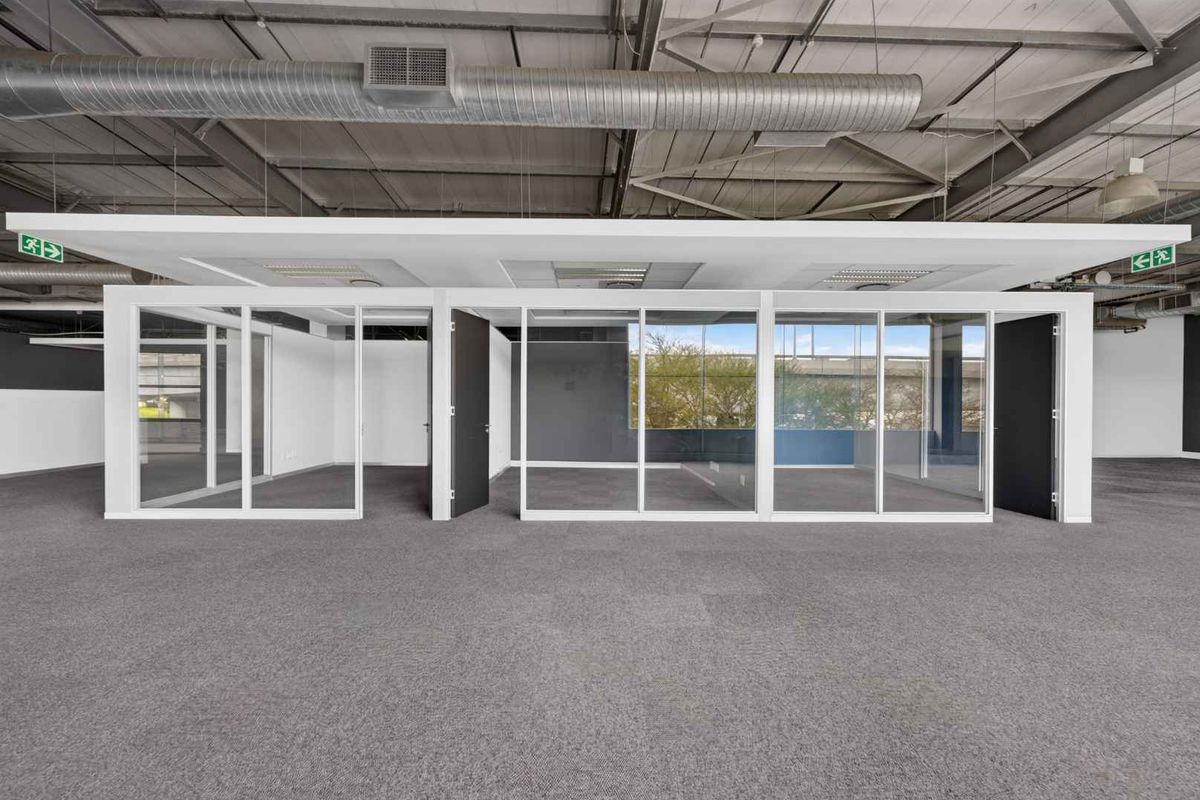 Modern open-plan office space featuring glass-partitioned private offices, exposed industrial ceiling ductwork, and neutral carpeting throughout.