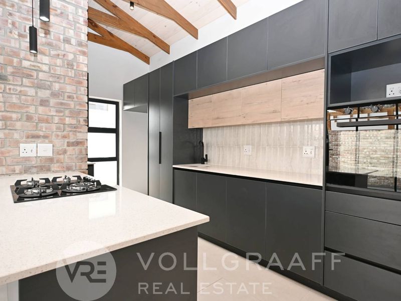 Modern kitchen featuring dark cabinetry, stone countertops, gas hob, built-in oven, and an exposed brick wall with timber ceiling beams.