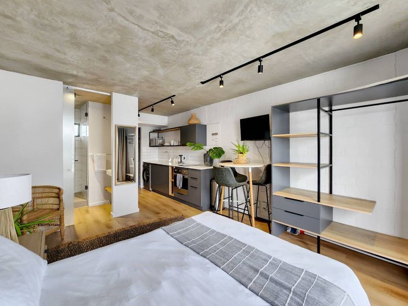 Modern open-plan studio apartment featuring an exposed concrete ceiling, sleek kitchen cabinetry with built-in appliances, timber flooring, and an en-suite walk-in shower.