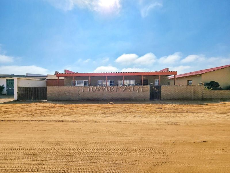 Front elevation of a single-story house featuring a covered patio, perimeter brick wall, and metal gate, situated on an unpaved sandy street.