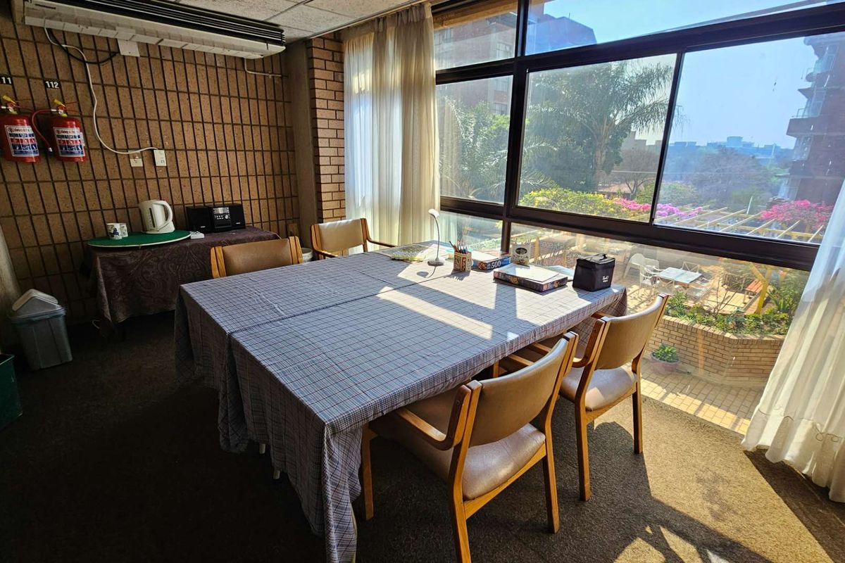 A communal living or dining area featuring large windows with natural light, brick feature walls, and carpeted flooring within a retirement village setting.