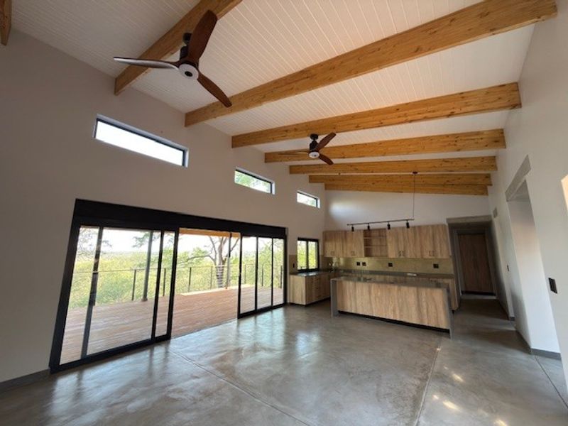 Spacious open-plan living and kitchen area featuring polished concrete floors, exposed timber beams, and large sliding doors opening onto a wooden deck.