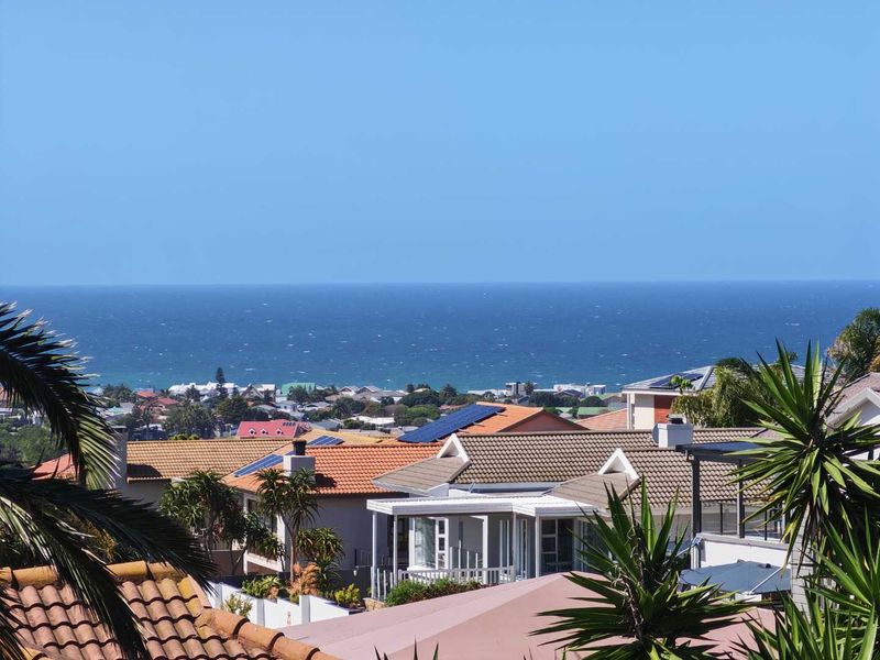 An elevated coastal view overlooking a residential neighborhood with tiled roofs and solar panels, extending out to the ocean horizon.