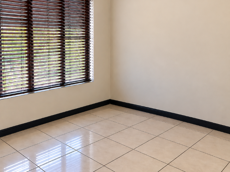 A bright, empty bedroom featuring neutral tiled flooring, dark skirting boards, and fitted venetian blinds.