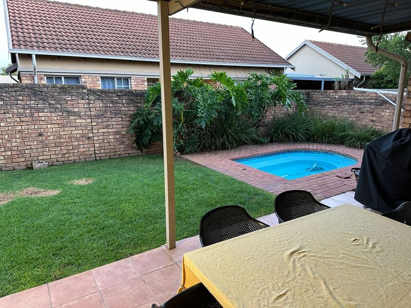 A covered patio area overlooking a private garden featuring a small swimming pool, brick boundary walls, and a lawn.