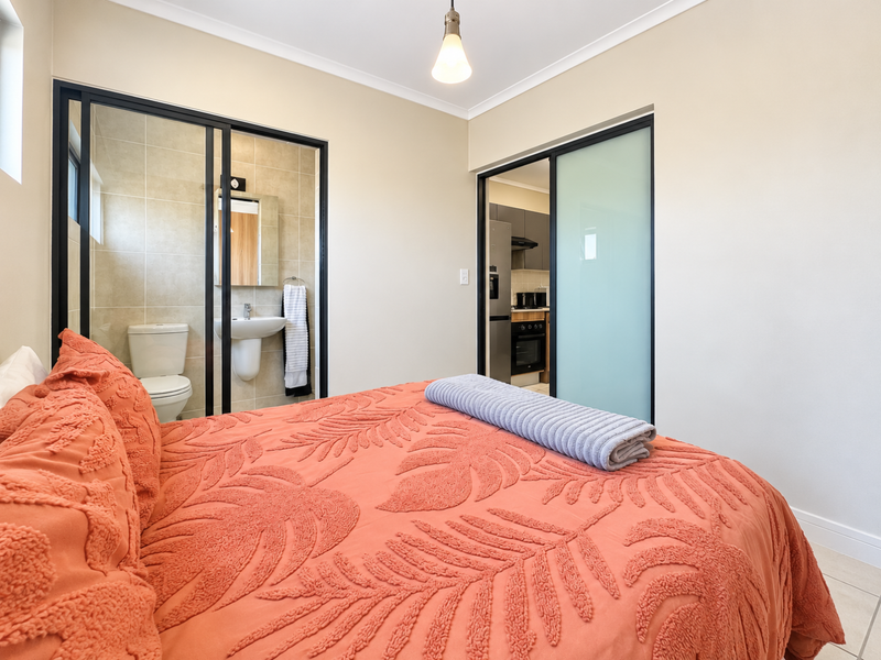 A well-maintained bedroom featuring sliding glass doors leading to an en-suite bathroom and adjacent kitchen area, with neutral walls and tiled flooring.