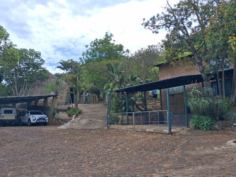 Front exterior view of the farm property featuring a cobblestone driveway, metal carports, and stone masonry building structures set within a landscaped garden.