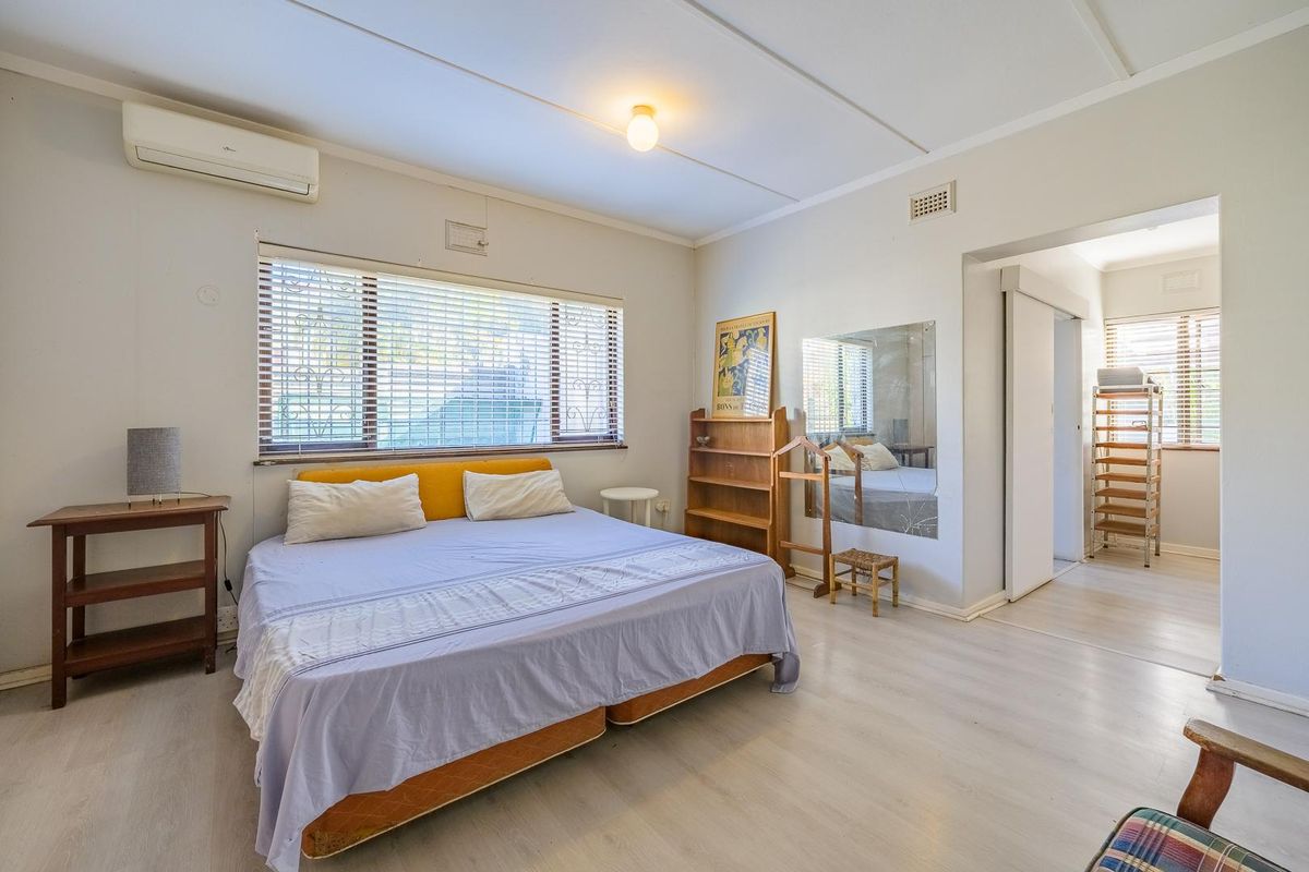 Spacious bedroom featuring laminate flooring, a wall-mounted split-system air conditioner, and large windows with venetian blinds, leading to an adjoining space.
