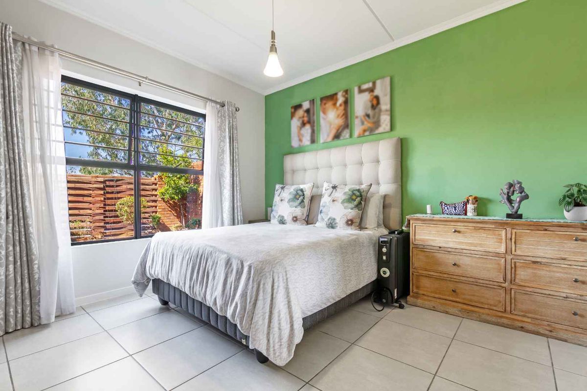 A well-lit bedroom featuring neutral tiled flooring, a large window with security bars, and a vibrant green accent wall.