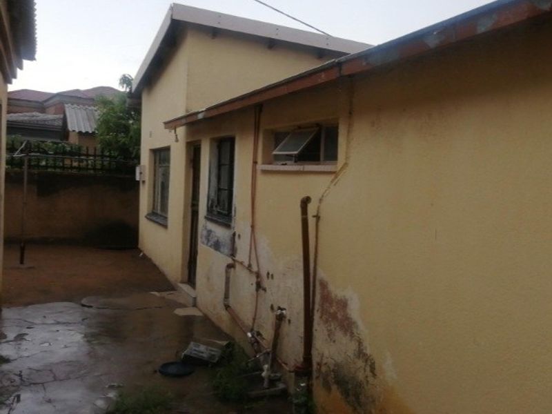 Rear exterior view of the property showing exposed plumbing, weathered wall finishes, and concrete paving requiring maintenance and repair.