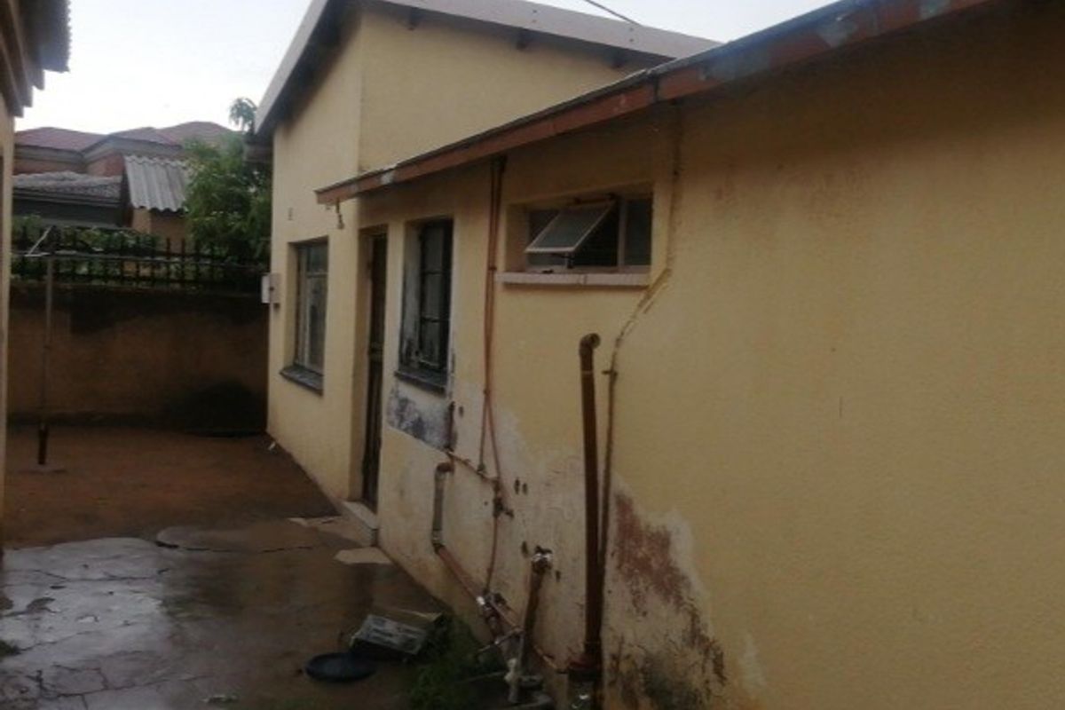 Rear exterior view of the property showing exposed plumbing, weathered wall finishes, and concrete paving requiring maintenance and repair.