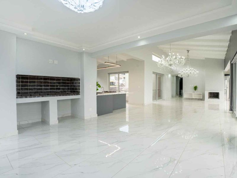 Spacious open-plan living area featuring polished marble-look tiles, elegant crystal chandeliers, a built-in braai, and seamless flow to the kitchen and outdoor space.