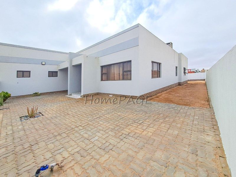 A modern single-story house exterior featuring a paved driveway, neutral facade, and aluminium window frames, set within a walled perimeter.