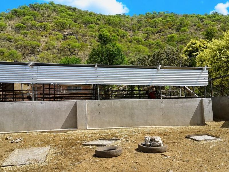 An outdoor livestock enclosure featuring a concrete perimeter wall, metal fencing, and a corrugated metal roof structure set against a natural mountain landscape.
