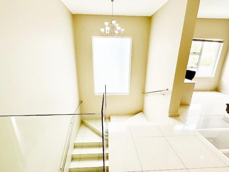A bright, modern hallway featuring a tiled staircase with a glass balustrade, stainless steel handrail, and a contemporary chandelier.
