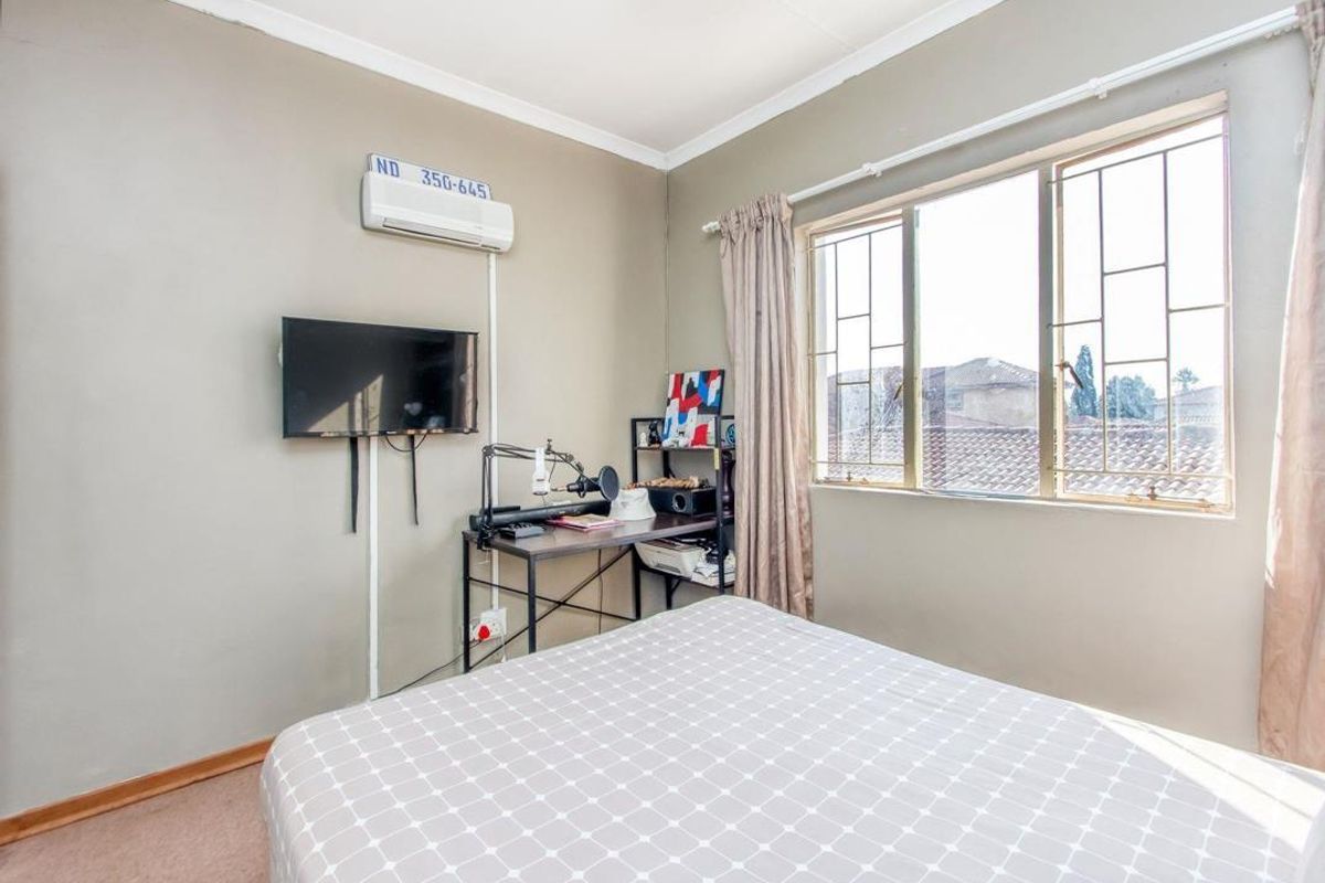 A functional bedroom featuring a wall-mounted air conditioning unit, security bars on the window, and neutral wall finishes.