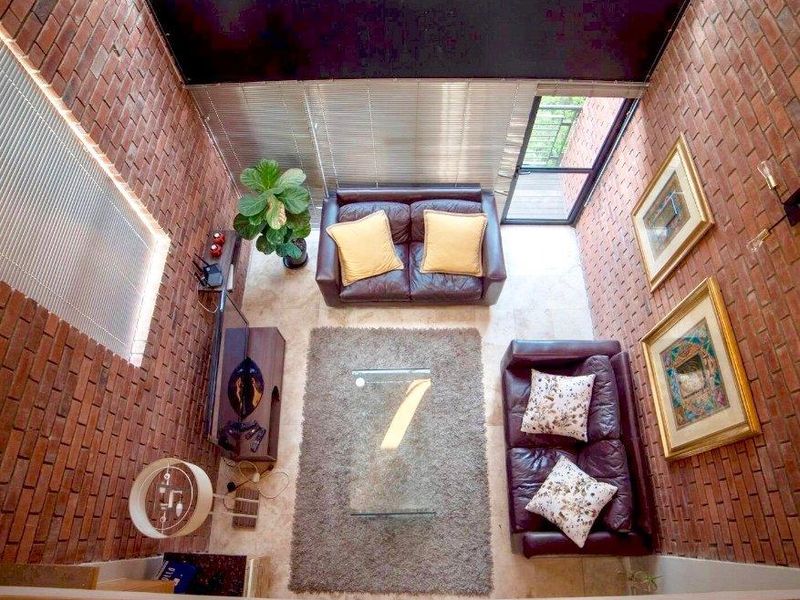 A double-volume living room featuring exposed brick walls, tiled flooring, and large windows with venetian blinds, providing access to an outdoor area.