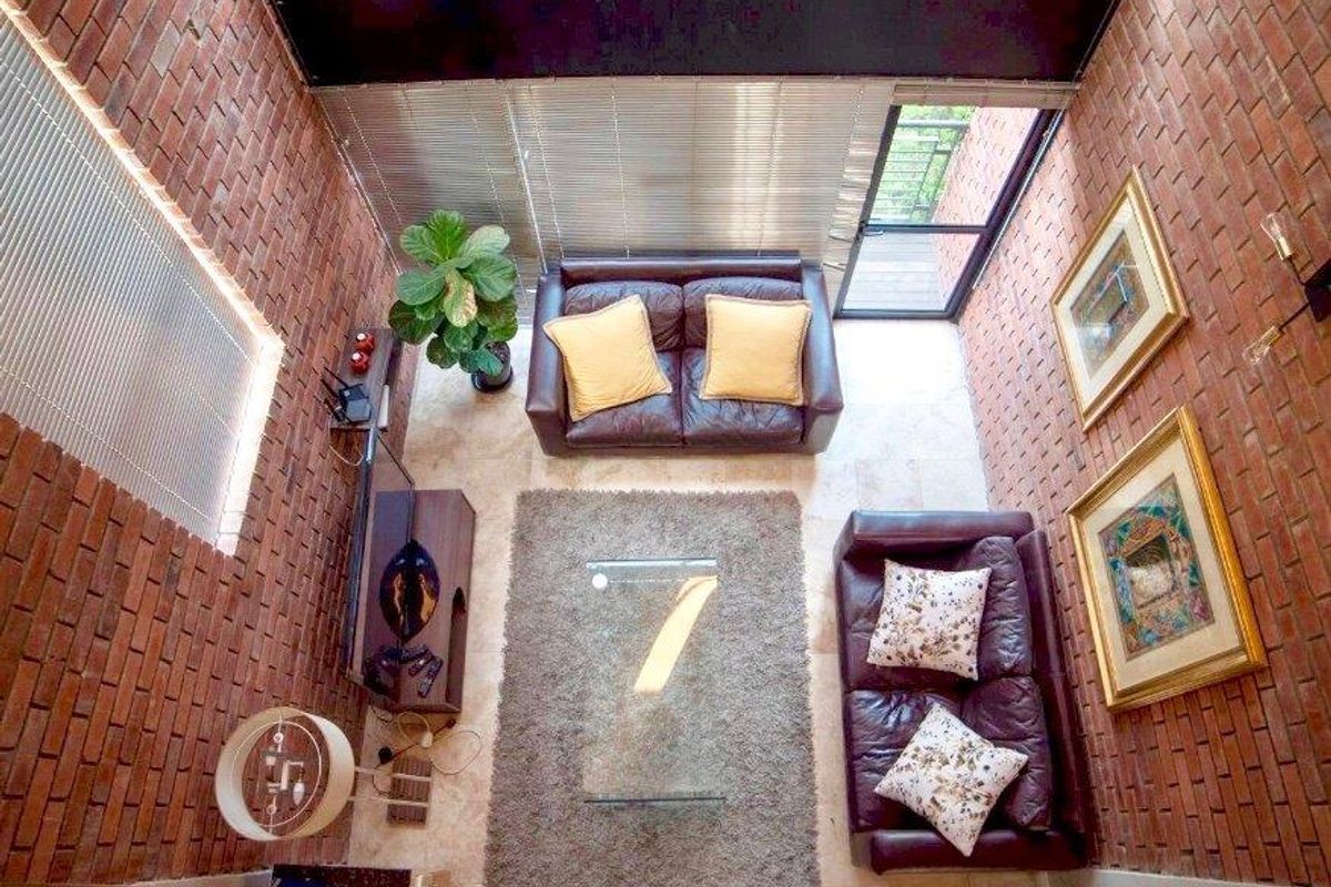A double-volume living room featuring exposed brick walls, tiled flooring, and large windows with venetian blinds, providing access to an outdoor area.