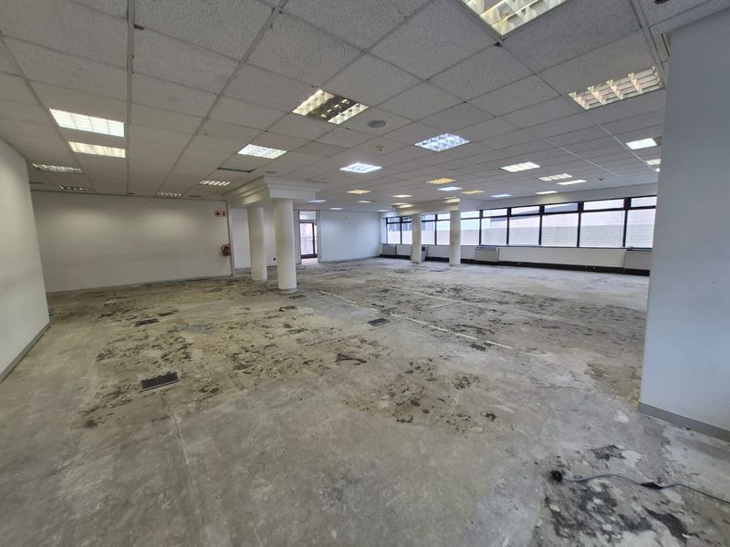 A large, vacant open-plan office space featuring a suspended ceiling, recessed lighting, structural columns, and large windows, currently requiring significant flooring repairs.