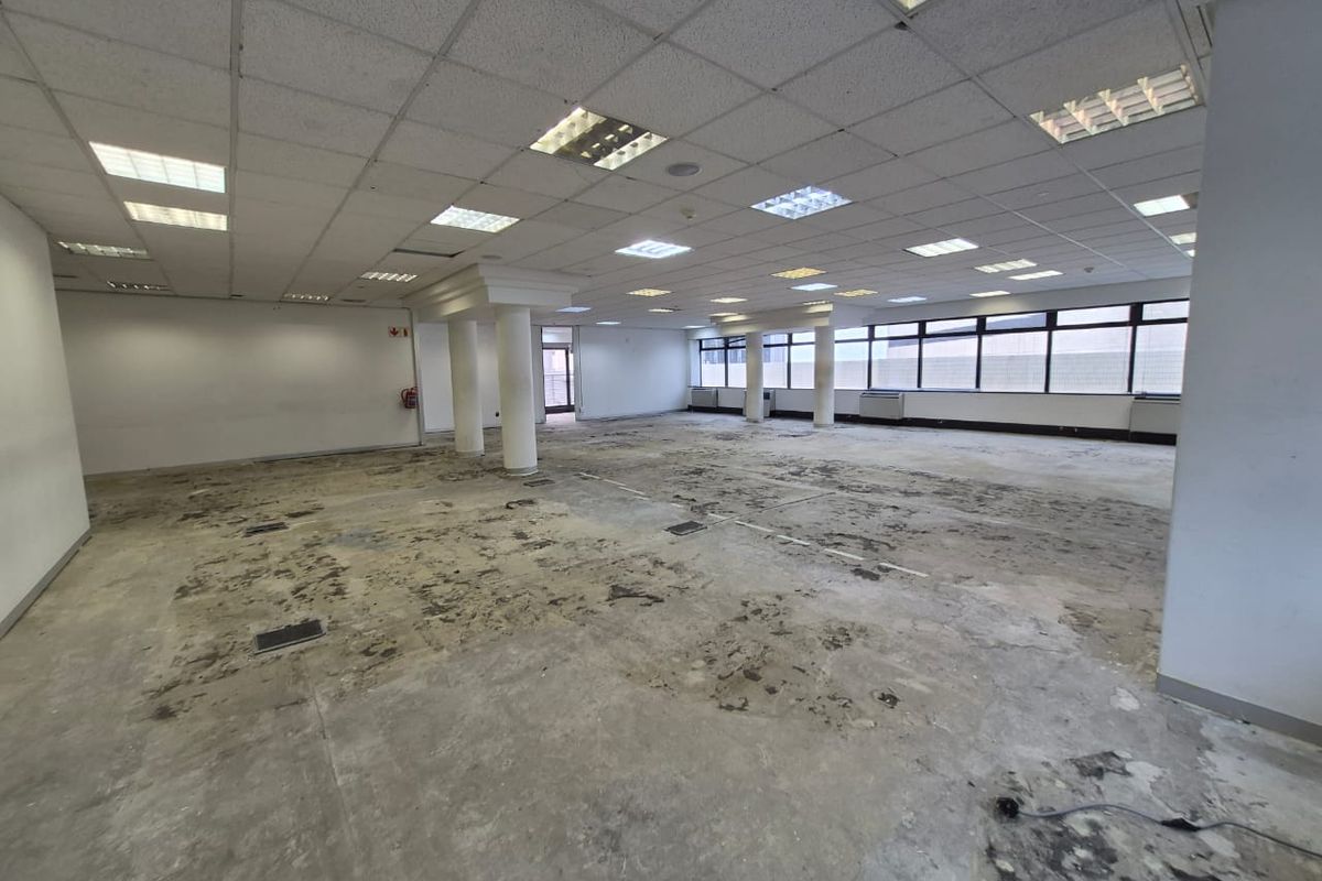 A large, vacant open-plan office space featuring a suspended ceiling, recessed lighting, structural columns, and large windows, currently requiring significant flooring repairs.