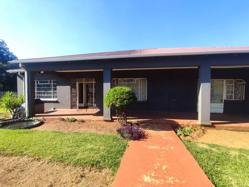Front elevation of a house featuring a covered patio, metal roofing, and security gates, set against a landscaped garden with a concrete walkway.