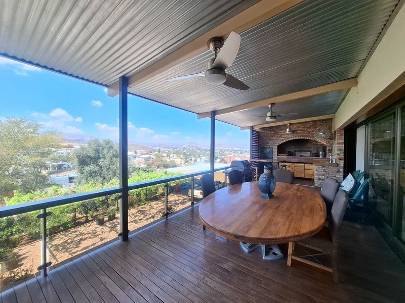 Spacious covered patio featuring wooden decking, a built-in brick braai, ceiling fans, and a glass balustrade overlooking the surrounding landscape.