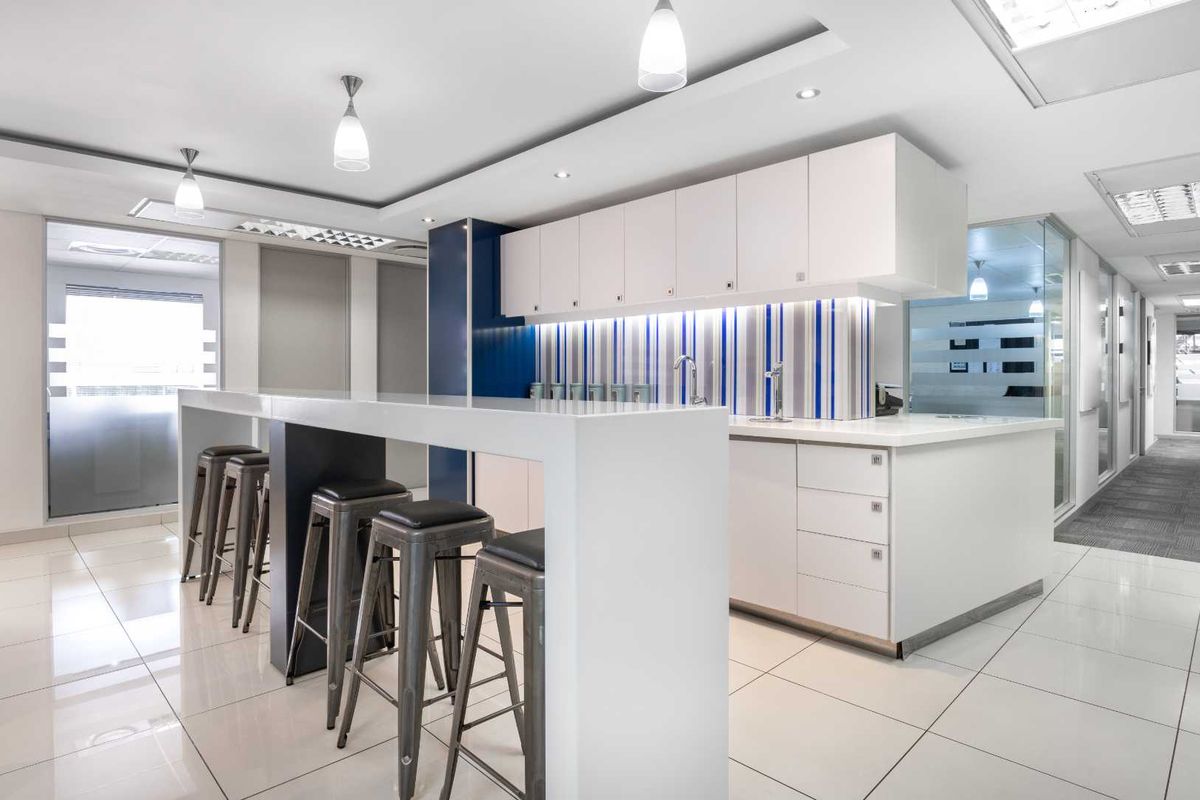 Modern office kitchenette featuring sleek white cabinetry, a breakfast bar, integrated sink, and contemporary lighting in a professional workspace.