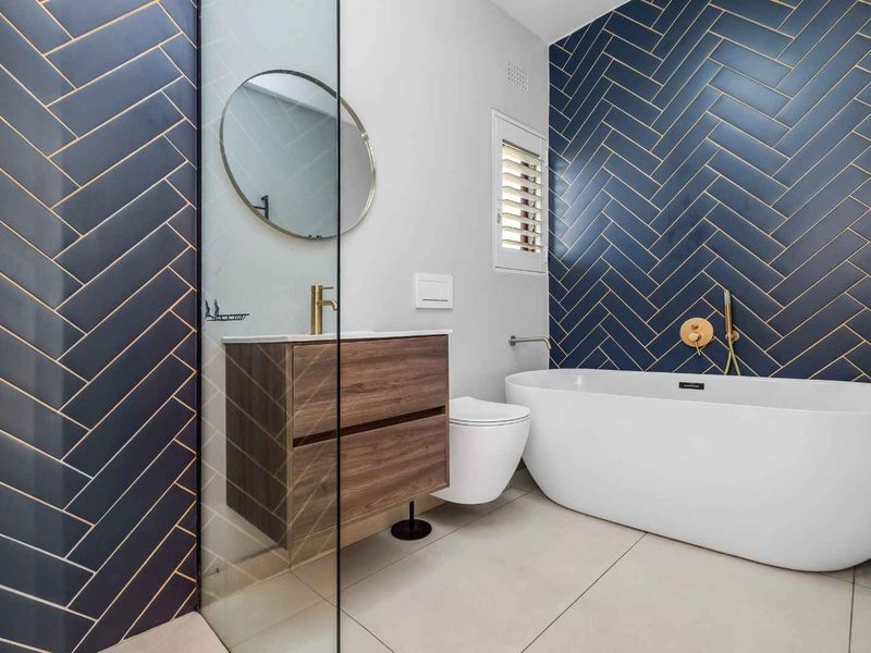 Modern bathroom featuring a freestanding bathtub, wall-hung vanity, wall-hung toilet, and striking navy blue herringbone wall tiles with brass accents.
