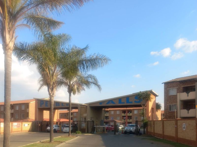 The entrance to Pebble Falls apartment complex featuring a secure gated access point, brick boundary walls, and established landscaping.