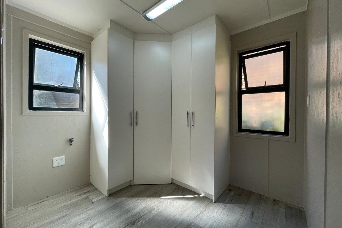 A bright bedroom featuring built-in white corner cupboards, modern wood-look laminate flooring, and dual windows providing natural light.