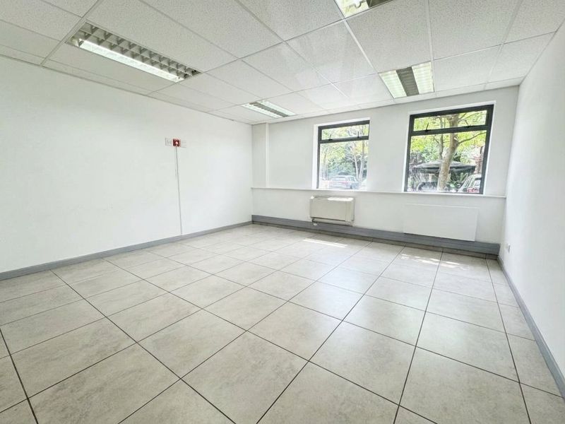 A bright, empty office space featuring tiled flooring, a suspended ceiling with recessed lighting, and wall-mounted HVAC unit with large windows.
