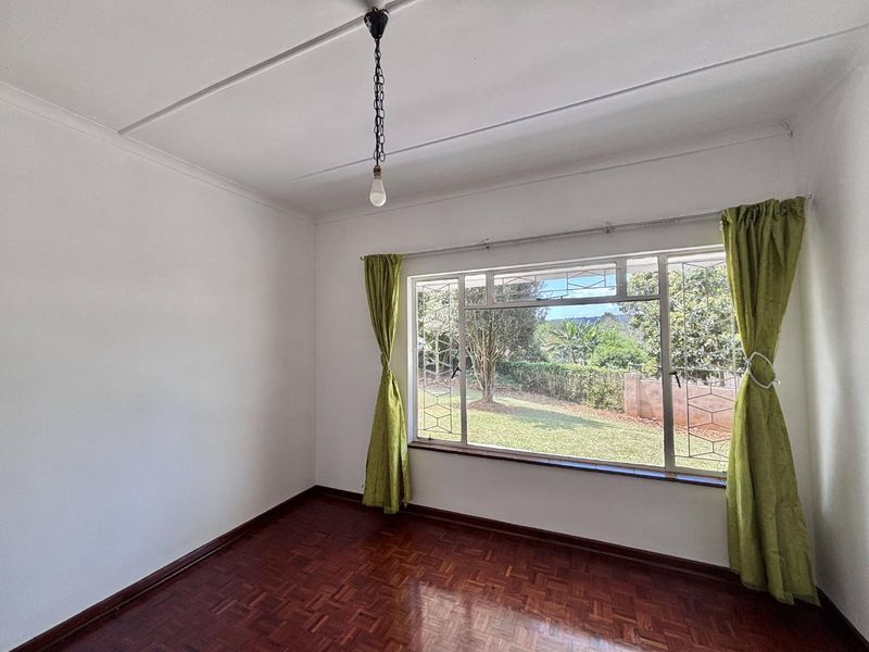 A bright, empty bedroom featuring classic parquet flooring, a large window with security bars, and a simple pendant light fixture.