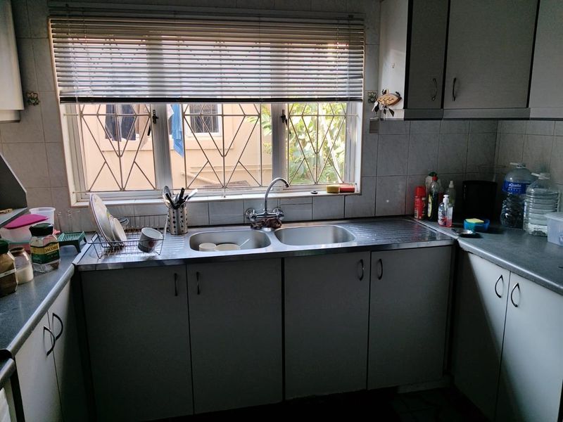 A functional kitchen featuring a double bowl stainless steel sink, laminate countertops, and built-in cabinetry with a tiled backsplash and window security bars.