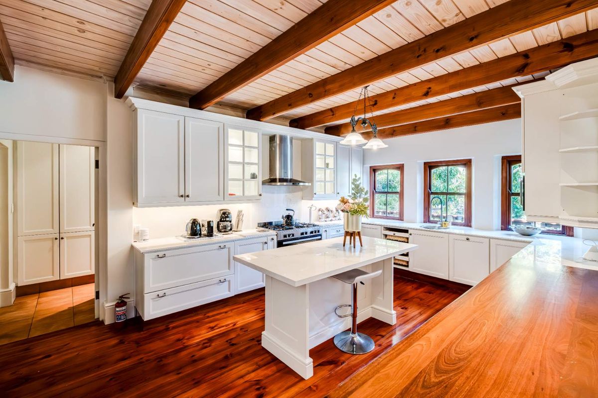 A well-appointed kitchen featuring white cabinetry, a central island with stone countertop, stainless steel extractor fan, and exposed timber ceiling beams.