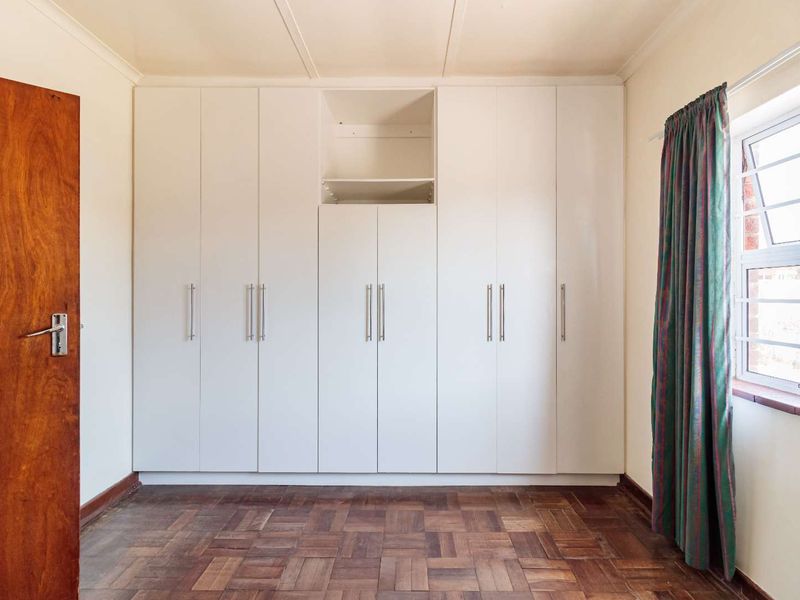 A bright, empty bedroom featuring classic parquet flooring, ample built-in cupboard storage, and a window with natural light.