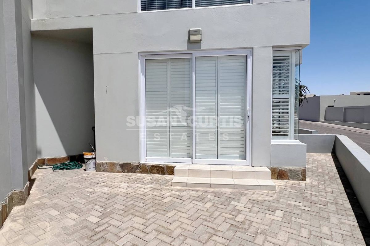 A paved rear patio area featuring sliding glass doors with white shutters, stone base detailing, and a clean exterior facade.